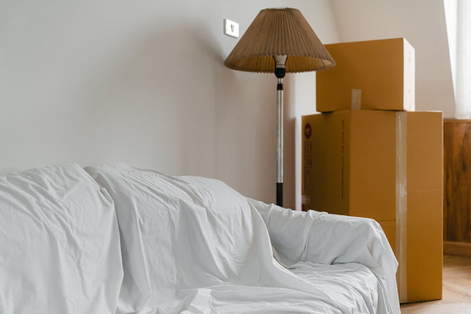 In a residential interior, a white fabric-covered sofa is positioned in front of a plain light-colored wall, with soft natural light illuminating the scene. To the right of the sofa, there are three cardboard boxes stacked vertically near a wooden piece of furniture, likely a cabinet or sideboard. A tall floor lamp with a brown pleated lampshade is placed beside the boxes, providing additional lighting. The boxes appear to be sealed with packing tape, suggesting they are prepared for transport or unpacking as part of a home relocation process. The setting shows a clean, organized space with a hardwood floor, typical of a living room prepared for furniture and packing items to be moved, illustrating a typical furniture transport and packing scenario in a house removal service, as handled by Man and Van Northwood.