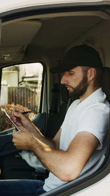 A man wearing a black cap and white shirt is standing indoors next to a storage area filled with stacked cardboard boxes of various sizes. He is holding a clipboard and a pen, appearing to take invent