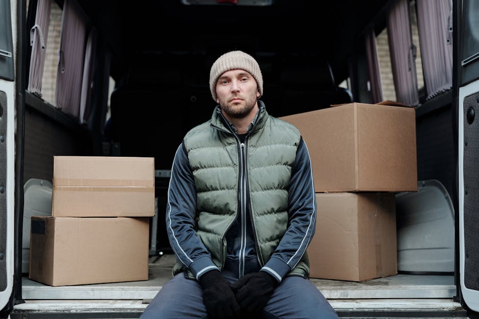 A man wearing a grey cap, black gloves, a navy blue jacket with a black padded vest, and black trousers is standing outside a residential property near a white moving van. He is leaning against the op