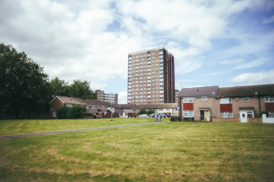 A residential area in Northwood featuring a large, open grassy field with a paved pathway running through it, flanked by a row of single-story houses with brick and beige exterior walls, some with white window frames and small front gardens. In the background, a tall, multi-storey residential building with a brown brick facade and numerous windows rises above the lower houses, under a partly cloudy sky with patches of blue. The scene is well-lit with natural daylight, demonstrating the surroundings often involved in home relocation and furniture transport processes. The image represents a typical suburban setting where house removals and packing and moving activities may take place, with the proximity of a potential loading area for a removal van or service vehicle. The presence of parked cars and the open space underscores the logistics involved in house removals, as managed by companies like Man and Van Northwood, within the context of local street loading regulations.