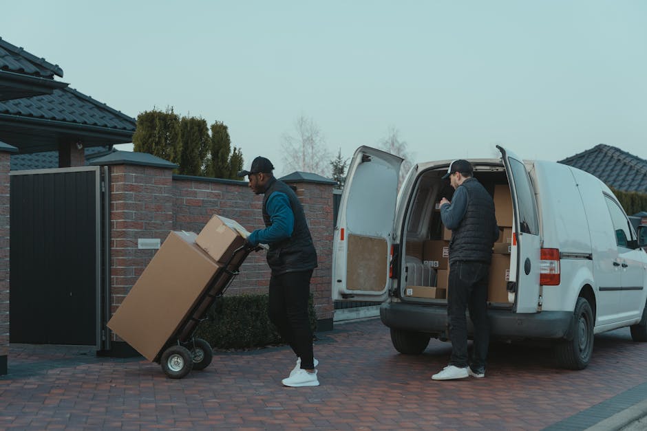Two men engaged in a home relocation process are seen loading cardboard boxes into a white panel van parked on a brick-paved driveway during daytime with overcast weather. One man, wearing a blue and black jacket, black trousers, white sneakers, and a cap, is pushing a black hand truck with a large cardboard box towards the open rear door of the van. The other man, dressed in a black puffer vest, dark trousers, and a cap, is standing inside the van, organizing or packing boxes on the vehicle's floor, which is lined with more cardboard boxes and packaging materials. The vehicle is positioned close to a brick wall with a black metal gate and surrounding fencing, and residential houses with tiled roofs and trees are visible in the background. The scene demonstrates a professional moving and packing process, emphasizing furniture transport, loading, and packing during a home relocation, with [COMPANY_NAME] providing the removals service.
