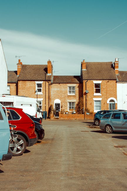 A wide view of a residential street showing a row of terraced brick houses with small front gardens, under a clear blue sky with a faint cloud and a contrail. In the foreground, a parking area features several parked vehicles, including a red SUV, a blue hatchback, and a grey car. The parking lot appears to be part of a home relocation process managed by Man and Van Northwood, as part of their removals services. Two individuals are visible near the front doors of the houses, possibly involved in packing or loading activities. The scene suggests a typical urban setting where furniture and boxes may be being prepared for transport, with the vehicles potentially used for furniture transport or home relocation. The environment indicates a daytime setting with natural lighting, and the overall context aligns with a professional moving operation involving loading, packing, and arranging of household items for a house removal in Northwood.