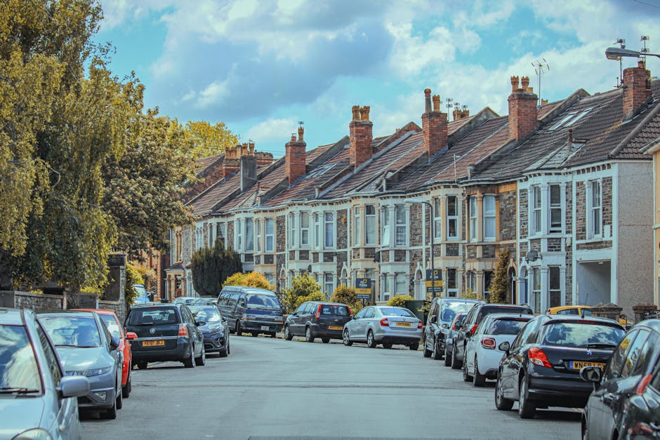 A residential street with a row of terraced houses featuring brick and white-painted facades, each with multiple chimneys and tiled roofs under a partly cloudy sky. Parked cars line both sides of the street, with some vehicles partially visible, including sedans, hatchbacks, and vans, arranged along the curb and on the pavement. A large tree with green foliage is situated on the left side, partially obscuring some houses, while the right side shows a clear view of house entrances and front gardens. The street appears to be part of a typical UK neighbourhood, suitable for house removals and relocation services. The scene illustrates the environment in which Man and Van Northwood conducts home relocation, involving careful loading and transport of household furniture, boxes, and appliances, often requiring the use of trolleys, straps, or blankets for safe furniture transport within tight urban spaces.