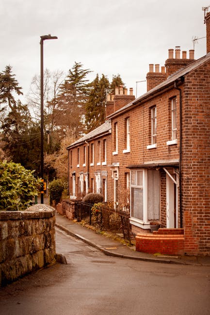 A narrow residential street in Northwood with a row of traditional red brick terraced houses featuring white window frames and clay tile roofs. The pavement is paved with concrete and lined with low brick walls and greenery, including bushes and small trees. A black lamppost is located on the sidewalk near the edge of the frame. The sky is overcast, creating diffuse lighting that highlights the details of the houses and the street scene. This setting provides a typical backdrop for home relocation or furniture transport activities carried out by Man and Van Northwood, especially when dealing with tight access areas in house removals, where careful planning of packing, loading, and vehicle positioning is essential for a smooth moving process.