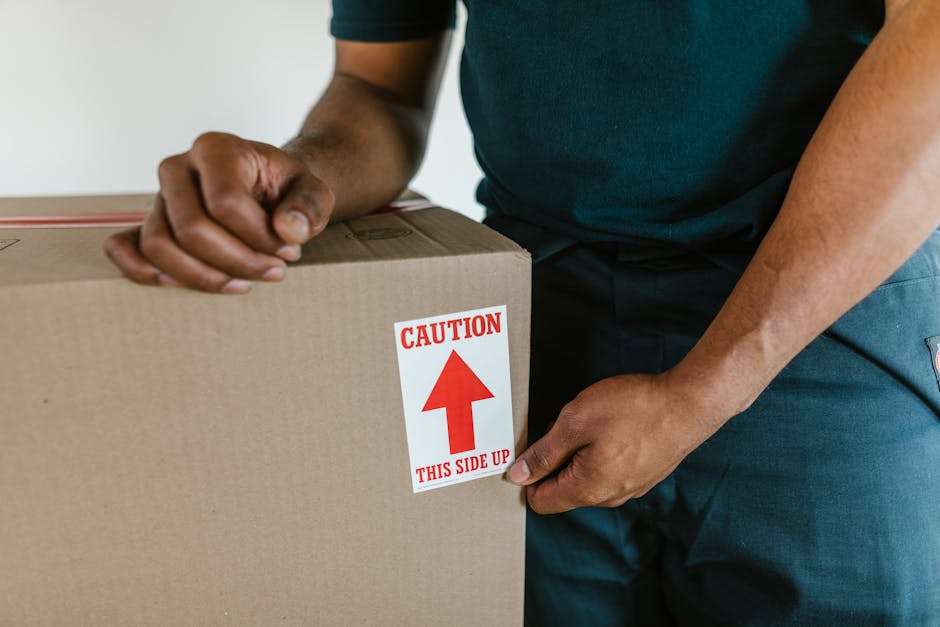 A person wearing a dark green shirt and blue work trousers is holding a large cardboard box marked with a red caution label that reads 'CAUTION THIS SIDE UP' and features an upward arrow, during a home relocation process. The individual is standing indoors on a plain, light-colored floor, positioned near a doorway or open space. The box is sealed with brown packing tape, indicating it contains fragile or important items for moving. The person's hands are gripping the box securely, with one hand on top and the other supporting it from underneath, suggesting they are in the process of lifting or loading the box into a moving vehicle or into a storage area as part of furniture transport or packing and moving activities. Behind the person's torso, part of a wooden door frame or wall is visible, and natural light illuminates the scene, highlighting the careful handling involved in the loading process. This image reflects professional removals services, such as those offered by Man and Van Northwood, that focus on delicate and efficient home removals, especially where careful packing and loading are required for tight access or narrow spaces during house relocations.