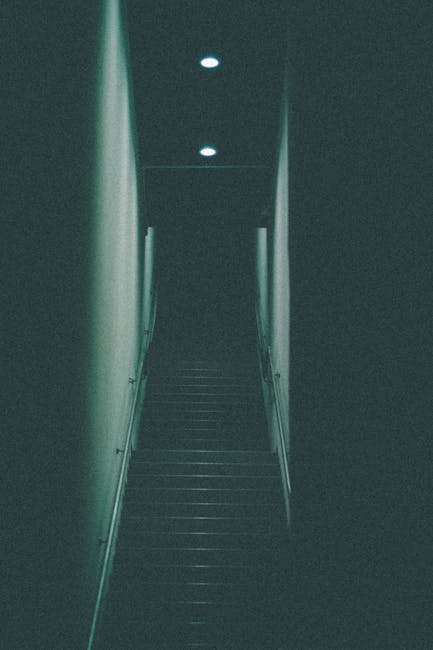 A narrow staircase inside a residential property illuminated by three small ceiling lights, with dark-colored steps and plain light-colored walls on either side. The staircase appears to lead to an upper floor, with a handrail visible on the right side. In the background, the top of the staircase is dimly lit, suggesting limited natural light. The scene depicts an interior space ready for a home relocation or moving process, with no additional furniture or items visible on the stairs or in the immediate vicinity. The image illustrates the challenge of navigating narrow staircases during furniture transport and packing during house removals, with [COMPANY_NAME], Man and Van Northwood, providing solutions for moving items through confined spaces like this.