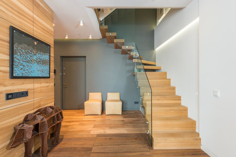 An interior view of a modern home with a wooden staircase featuring glass balustrades leading to an upper floor, positioned next to a grey wall with minimalistic furniture. The foreground shows a polished wooden floor with two small beige upholstered chairs placed against the wall near a grey door. To the left, a flat-screen television is mounted on a wooden-paneled wall, and below it, there are two decorative wooden animal sculptures. The lighting includes recessed ceiling lights, illuminating the space evenly. This setting appears to be part of a home relocation process, with the staircase and open layout facilitating efficient furniture transport and packing for a house move, as coordinated by Man and Van Northwood.