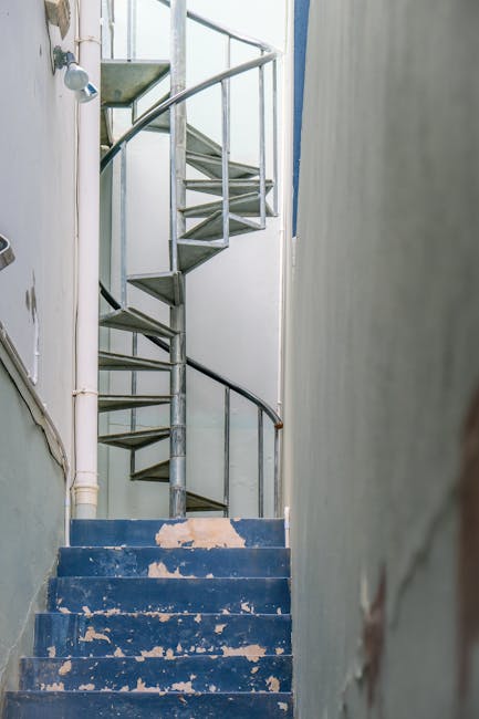 A narrow staircase inside a residential property illuminated by three small ceiling lights, with dark-colored steps and plain light-colored walls on either side. The staircase appears to lead to an upper floor, with a handrail visible on the right side. In the background, the top of the staircase is dimly lit, suggesting limited natural light. The scene depicts an interior space ready for a home relocation or moving process, with no additional furniture or items visible on the stairs or in the immediate vicinity. The image illustrates the challenge of navigating narrow staircases during furniture transport and packing during house removals, with [COMPANY_NAME], Man and Van Northwood, providing solutions for moving items through confined spaces like this.