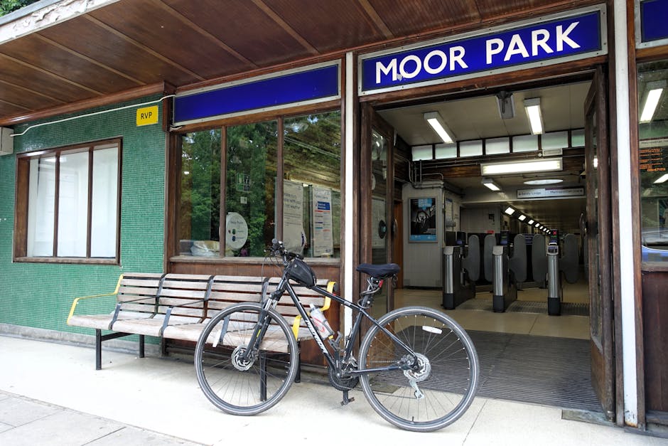 Exterior view of Moor Park station entrance showing a wooden and glass facade with a blue sign reading 'MOOR PARK' above the ticket barriers. In front of the station, a black bicycle with a water bottle attached to its frame is parked against a wooden bench with metal armrests, positioned on the pavement just outside the entrance. The pavement is light grey concrete, and the station entrance is flanked by large glass windows revealing the interior ticket barriers and turnstiles, which have black waist-height barriers and overhead lighting. The interior lighting creates a well-lit environment inside, while the exterior includes a green tiled wall with a small yellow 'RVP' sign near the window. The scene captures a typical setting for home relocation or furniture transport, with the station providing access for moving logistics, and the bicycle indicating urban accessibility. The overall environment appears calm and functional, suitable for facilitating a house removals service like those provided by Man and Van Northwood.