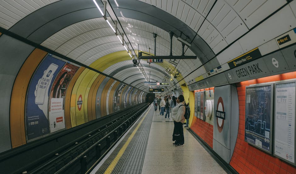 In the underground Green Park station at Northwood, London, a group of commuters is standing on the tiled platform near the edge, preparing to board a train. The station's curved tunnel walls are decorated with colourful advertising posters, including a prominent one on the left side featuring a white product on a blue background. The platform is equipped with digital display boards showing journey information, and safety markings are visible along the edge. Overhead, lighting strips illuminate the area, and structural support beams are suspended from the arched ceiling. The scene captures a typical busy moment during peak hours, with passengers carrying backpacks and luggage, some looking towards the tunnel entrance while others wait attentively for the train's arrival. Occasionally, staff or commuters are seen moving about, reflecting the bustling environment of a London underground station, which could be relevant for logistics discussions on house removals and furniture transport, especially in urban underground contexts.