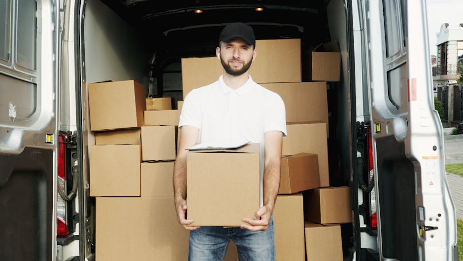 A man with a beard, dressed in a white shirt and black cap, is standing inside the open rear of a black moving van. He is holding a medium-sized cardboard box with both hands, positioned in front of h