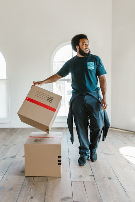 A man with dark curly hair and a beard is inside a bright, empty room with white walls and wooden flooring, engaged in a furniture transport task as part of a home relocation. He is wearing a navy blue T-shirt with a logo on the chest and dark trousers. The man is holding a cardboard box secured with red packing tape, lifting it in one hand while standing next to two stacked cardboard boxes on the floor. Behind him, there is a large arched window allowing natural light into the space, casting soft shadows on the floor. The room appears to be prepared for moving activities, with minimal furniture present, emphasizing the focus on packing and loading. This scene exemplifies the careful handling and organized approach essential for successful house removals, a service provided by Man and Van Northwood, supporting clients with efficient furniture transport and packing during their move.