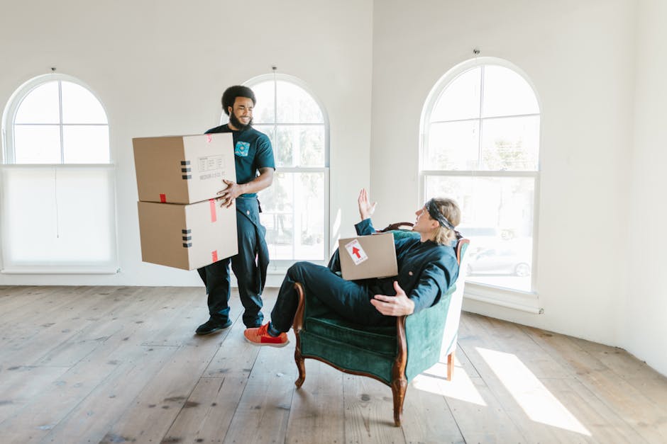 A man dressed in casual dark clothing and red shoes is seen carrying two medium-sized cardboard boxes, secured with red and black tape, through a bright, spacious room with white walls and large arched windows, which let in natural daylight. Inside the room, a woman in a dark jacket and glasses is seated on a vintage green armchair, holding a smaller cardboard box with a red arrow label, and raising her hand in a gesture of communication or greeting while looking at the man. The floor is made of light wooden planks, and the area appears to be in the process of a home relocation, with the boxes prepared for transport or unpacking. This scene captures a practical moment during packing and moving, illustrating the physical effort involved in furniture transport and the coordination typical of professional removals, possibly by Man and Van Northwood, as part of their house removal services.